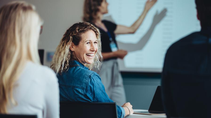 Personen sitzen in einer Präsentationssituation. Eine Frau dreht sich lachend zu einer anderen Person um.